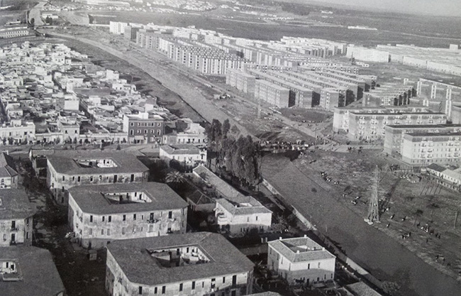 Fotografía aérea en blanco y negro de un barrio en desarrollo, con bloques de viviendas en construcción y áreas urbanas en expansión. Se observa el cauce del arroyo Tamarguillo y distintas zonas residenciales como Ciudad Jardín y Los Pajaritos en Sevilla.